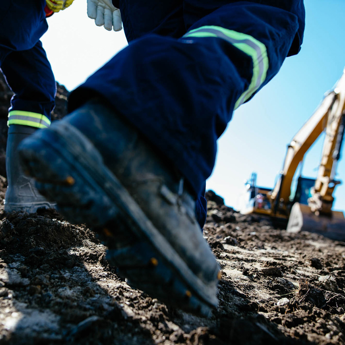 En premier-plan, les bottes de deux travailleurs dans la boue. En arrière-plan flou, de la machinerie lourde.