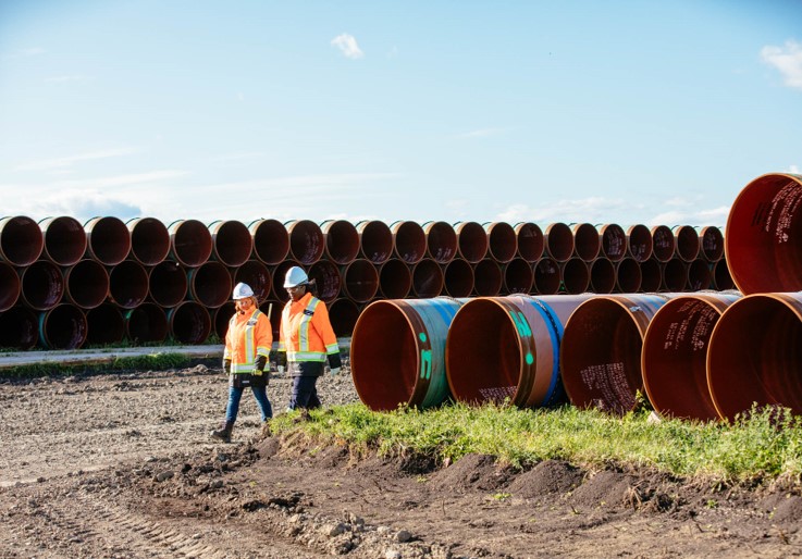 Two CER staff members wearing orange safety vests, white helmets, and protective gear walk along a dirt path beside a large stack of rust-coloured metal pipes arranged in horizontal rows. 
