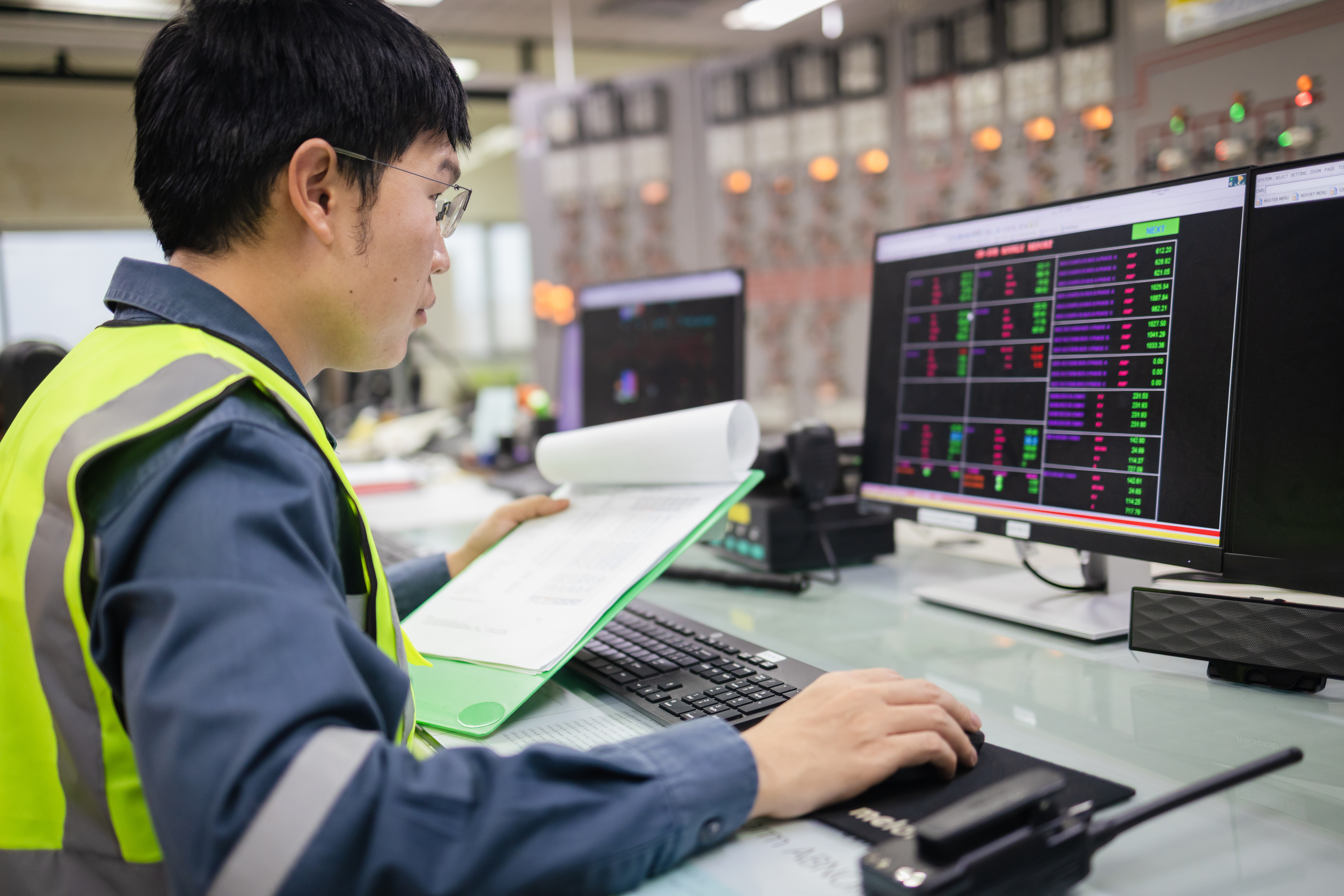 Vue latérale d'un ingénieur portant un gilet de sécurité et surveillant le réseau électrique sur deux écrans.