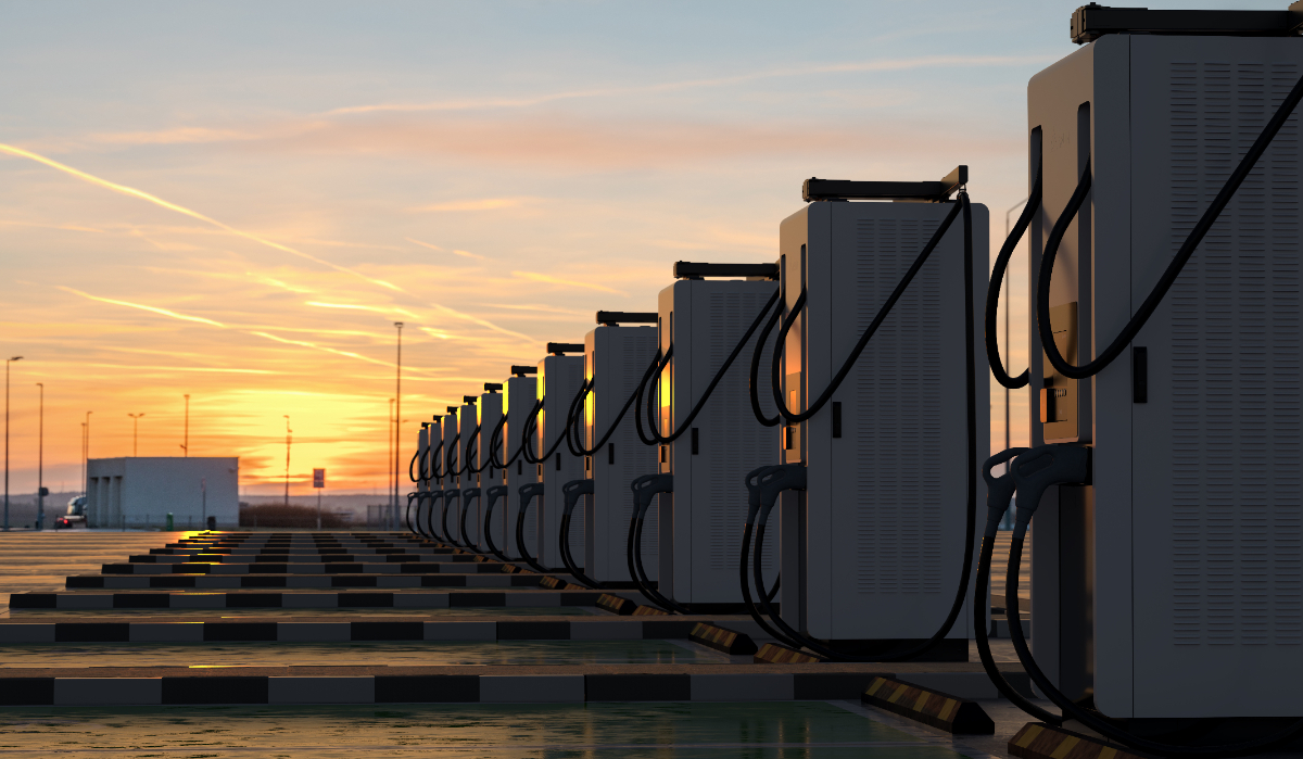 Plusieurs bornes de recharge rapide pour véhicules électriques alignées dans un stationnement, avec le soleil qui se couche derrière elles.
