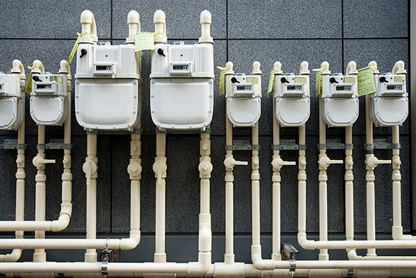 Compteurs blancs en succession contre un mur de briques grises d’un immeuble industriel reliés à des canalisations de gaz.