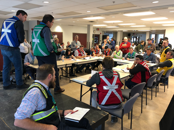 A group of staff in a room sitting down and listening to two individuals at the front. They are being briefed during an emergency exercise.